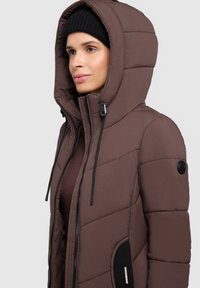 Jachetă puf galbenă cu glugă mare, închidere cu fermoar și şnururi negre. Prezintă cusături geometrice și un buzunar lateral cu branding.