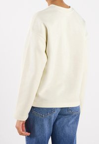 Persona che indossa una felpa a maniche lunghe beige semplice e jeans in denim blu, vista da dietro su uno sfondo bianco.