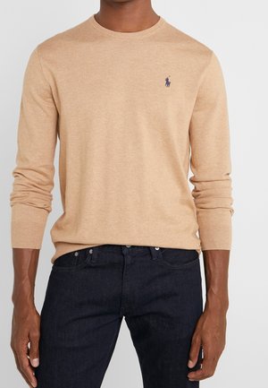 Mies, joka pukeutunut vaaleanruskeaan pitkähihaiseen neulepaitaan, jossa on pieni tummansininen Polo Player -logo rinnassa, yhdistetty tummansinisiin farkkuihin.