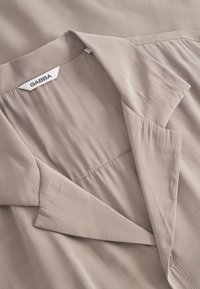 Beige blouse met kraag en zachte textuur, voorzien van een gevouwen halslijn en geplooide details. Label zichtbaar, ontwerp richt zich op eenvoud en elegantie.