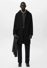 Abrigo de lana negro con capucha, cierre de botones y dos bolsillos. Lleva puesto un suéter gris y pantalones negros, sosteniendo una bufanda gris.