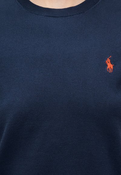 Μπλε σκούρο πλεκτό πουλόβερ με στρογγυλή λαιμόκοψη, με κεντημένο πορτοκαλί λογότυπο στο αριστερό στήθος. Ομαλή υφή, χωρίς ορατά σχέδια.