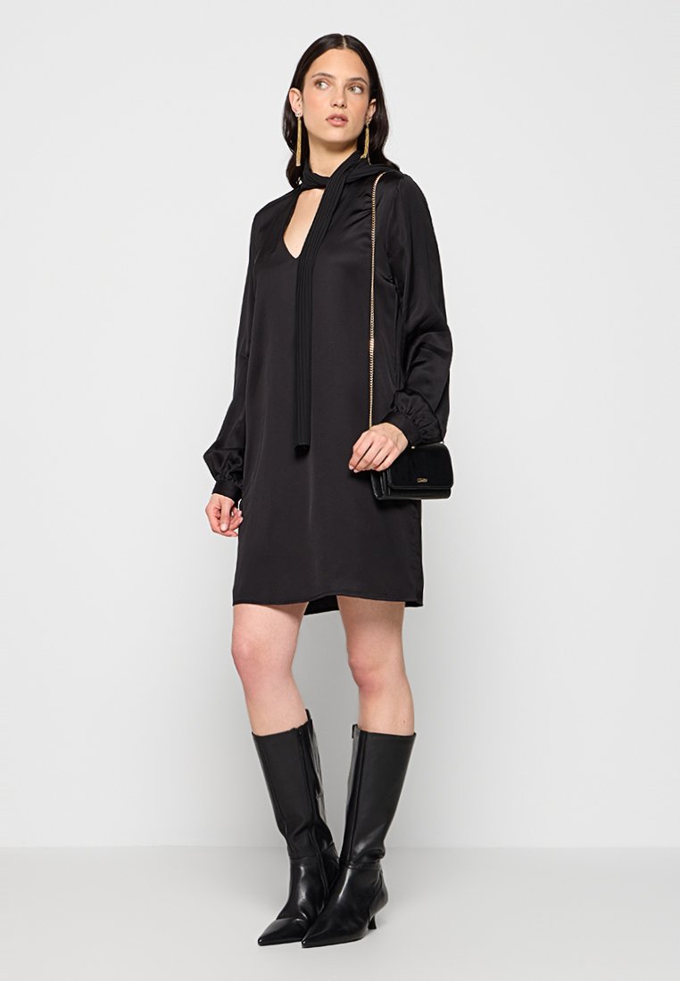 Abito in satin nero con scollatura a oblò, maniche lunghe a palloncino e dettagli plissettati. Abbinato a stivali neri al ginocchio e a una borsetta piccola.