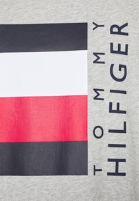 Tejido gris con un logotipo rectangular que presenta una franja superior azul marino, una franja media blanca y una franja inferior roja, junto al texto "TOMMY HILFIGER".