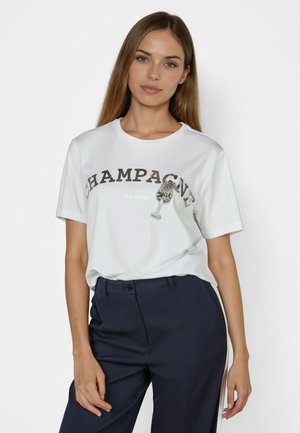 Young woman with long hair wears white T-shirt reading "CHAMPAGNE" with embellished champagne glass, paired with dark trousers.