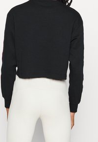 Korte zwarte sweatshirt met lange mouwen. Heeft een ronde halslijn en lichte ribbeling bij de boorden. Gedragen over lichtgekleurde leggings.