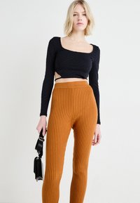 Zwarte lange mouwen crop top met een gestructureerd patroon, gecombineerd met ribbelige oranje hoge taillebroeken. Model houdt een klein zwart tasje vast.