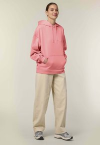 Felpa rosa con tasca a marsupio e cappuccio con cordino, abbinata a pantaloni beige a gamba larga e sneakers grigie. Sfondo semplice.