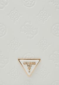 Hvid tekstureret overflade med en præget mønster. Gyldent trekantet logo med "GUESS USA" og et spørgsmålstegn i midten.