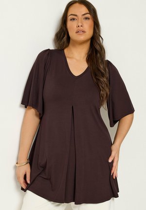 Mujer con cabello largo y castaño, vestida con una túnica suelta de color marrón oscuro con cuello en V y mangas cortas y anchas, de pie sobre un fondo blanco.