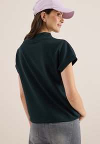 Mörkgrön pikétröja med upprullade ärmar, tillverkad av mjukt tyg. Bärs tillsammans med en ljuslila keps. Grå jeans syns vid den nedre kanten.