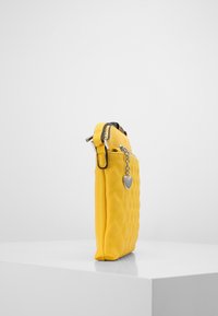 Kulttuurattu keltainen käsilaukku, jonka sivussa on hopeinen sydänkoru. Varustettu vetoketjulla ja teksturoituneella pinnalla. Kompakti suorakulmainen muoto.