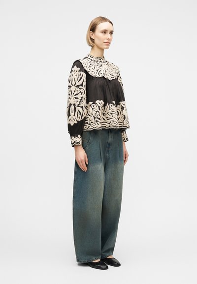 Femme debout portant un chemisier noir avec une grande broderie florale blanche, un jean large bleu et des ballerines noires, légèrement tournée vers la droite.