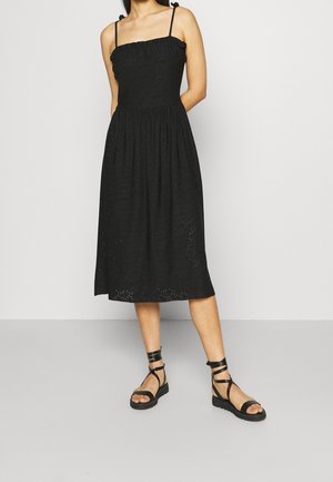 Frau trägt ein ärmelloses schwarzes Midikleid mit Bindeträgern und schwarze flache Sandalen mit Riemchen, steht vor einem schlichten hellen Hintergrund.