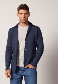 Navyblauer Cardigan aus strukturiertem Strickstoff, mit einem Schalkragen, vorderen Knöpfen und zwei Taschen. Getragen über einem leichten Pullover.