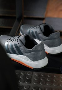 Zapatillas deportivas grises con parte superior de malla, acentos azules y una mediasuela blanca. Presentan una suela exterior texturizada con una sección naranja para mayor agarre.