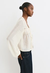 Helle, cremefarbene Bluse mit Puffärmeln, strukturiertem Stoff und einer Schleife vorne. Kombiniert mit dunklen Jeans.