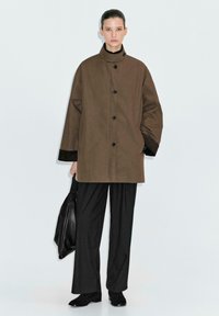 Cappotto marrone oversize con colletto alto, bottoni neri e polsini in velluto nero; abbinato a pantaloni neri a righe e una borsa nera.