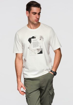 Junger Mann in weißem T-Shirt mit Grafik und olivgrünen Cargohosen, der nach unten schaut, eine Hand in der Tasche und eine Armbanduhr trägt.