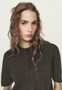 Brązowa koszulka polo z krótkimi rękawami, wykonana z dzianiny warkoczowej, z kołnierzykiem i trzema czarnymi guzikami z przodu, o fakturze miękkiej i przyjemnej w dotyku.