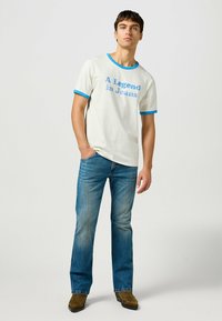 Camiseta de algodón blanca con cuello y mangas acanaladas azules, con "Una Leyenda en Jeans" impreso en azul en la parte delantera. Llevada con jeans azules.