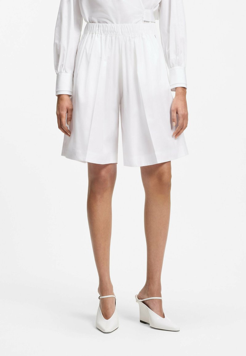 Femme portant un short blanc ample taille haute jusqu’aux genoux, une chemise blanche à manches longues et des mules blanches à talons avec brides à la cheville.