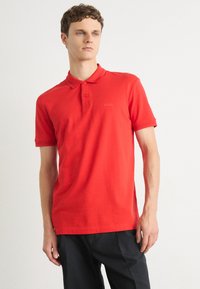Joven con una camisa polo roja con el logo "BOSS" y pantalones negros, de pie contra un fondo blanco liso.