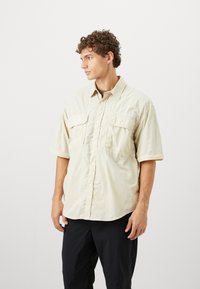 Camicia a maniche corte con bottoni in tessuto beige chiaro, due taschini sul petto, colletto a punta e vestibilità rilassata.