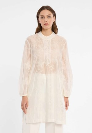 Vrouw die een doorschijnende, lange mouwen, crèmekleurige tuniek met bloemrijke borduurwerk en een hoge kraag draagt, staand tegen een effen witte achtergrond.