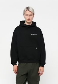 Zwarte hoodie met een voorzak en de tekst "VERSPEEL DE CULTUUR NIET". Gemaakt van zachte stof, met ribbelboorden en zoom.