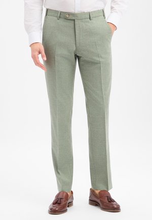 Mann trägt hellgrüne Anzughose, weißes Hemd und braune Ledertasselloafer, steht mit einer Hand in der Hosentasche.