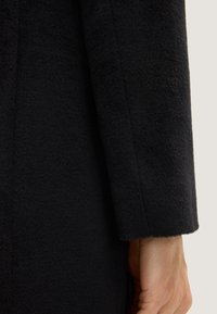 Zwart wollen stoffen jasje met een zachte textuur, met een gladde mouwrand en een close-up van een hand geklemd aan de zijkant.