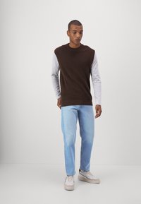 Maglione senza maniche marrone sopra una maglietta grigia a maniche lunghe, abbinato a jeans dritti blu chiaro e sneakers bianche. Design semplice e casual.