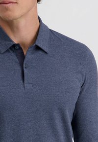 Mann in einem dunkelblauen Langarm-Polohemd mit zwei geöffneten Knöpfen vor einem schlichten hellen Hintergrund.