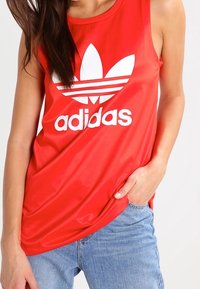 Röd ärmlös Adidas tanktop med en stor vit logotyp och tre horisontella ränder över bröstet, tillverkad av mjukt, lättviktsmaterial.