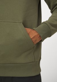 Olijfgroen sweatshirt met een voorzak, ribbelmanchetten en -zoom. De stof lijkt glad met een consistente afwerking en minimale stikdetails.