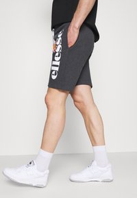 Shorts en coton gris avec poches latérales, arborant un grand logo blanc sur la jambe gauche. Portés avec des baskets blanches et des chaussettes blanches montant à la cheville.