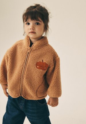 Bambino piccolo con capelli scuri che indossa una giacca in pile beige con una toppa zucca e jeans blu, in piedi contro uno sfondo neutro.