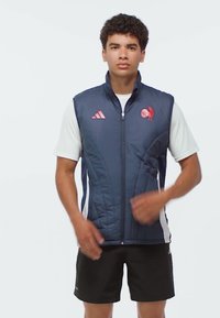 Junger Mann trägt eine marineblaue, ärmellose Steppweste über einem weißen Kurzarmhemd und schwarzen Shorts, steht vor einem schlichten hellen Hintergrund.