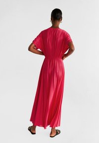 Robe maxi en tissu rose vif, avec un design plissé, des manches courtes amples et une taille élastique, accompagnée de sandales noires plates.