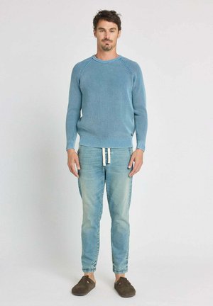 Pull bleu clair en tricot avec col et poignets côtelés, associé à un jean bleu délavé et des chaussures marron sans lacets. Design simple et décontracté.