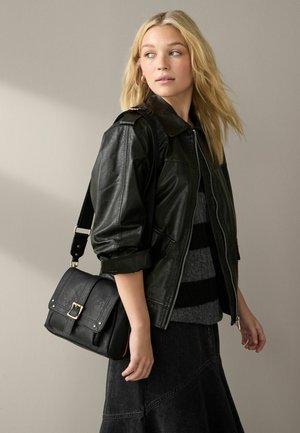 Giacca in pelle nera con zip frontale, maniche arrotolate, abbinata a una borsa a tracolla nera con fibbia e cinturino. Maglione grigio a righe sotto.