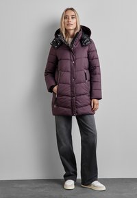 Lila pufferväst med en glansig svart huva, med ett quiltat mönster, dragkedjefickor och en medellång passform, tillsammans med mörka jeans och sneakers.