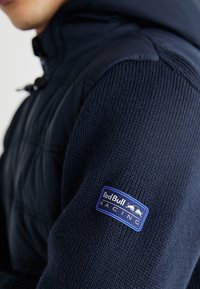 Granatowa kurtka z pikowanym korpusem i teksturowanymi, dzianinowymi rękawami, ozdobiona naszywką "Red Bull Racing" w niebiesko-białych kolorach na ramieniu.