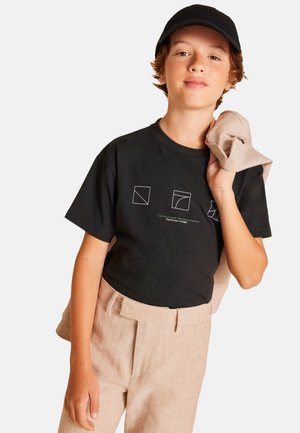 T-shirt nera in cotone con stampa geometrica bianca e testo, abbinata a pantaloni in lino beige chiaro. Un morbido maglione beige è appoggiato su un braccio.