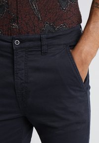 Tummansininen puuvillasekoitehousut, joissa on tasainen etuosa, sivutaskut ja sileä rakenne. Varusteena nappisulku ja vyölenkit.