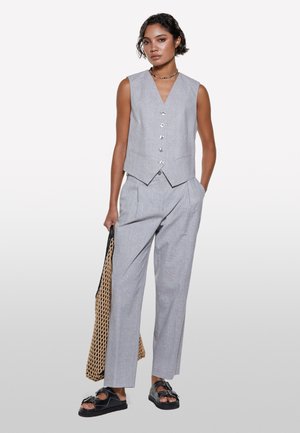 Gilet gris ajusté avec un col en V et cinq boutons, associé à un pantalon gris plissé taille haute. Le mannequin tient un sac en filet et porte des sandales.