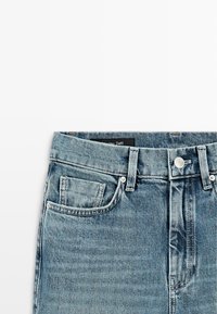 Lichtblauwe denim jeans met knopen aan de voorkant, riemlussen, voorzak en een zichtbaar merkje aan de binnenkant van de tailleband tegen een witte achtergrond.