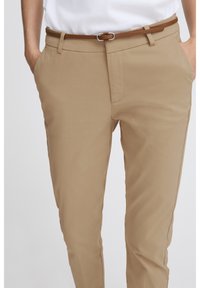 Pantalon tailleur beige à texture lisse, doté d'une ceinture marron avec une boucle métallique. Coupe moderne avec poches latérales et taille nette.