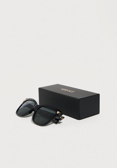 Μαύρα υπερμεγέθη γυαλιά ηλίου με σκοτεινούς φακούς δίπλα σε μαύρο κουτί με την επωνυμία Versace σε απλό λευκό φόντο.